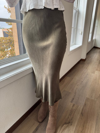 Person wearing a green skirt and beige boots standing in a room with wooden flooring and large windows.