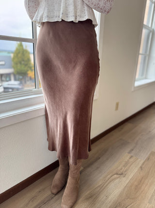Person wearing a brown skirt and beige boots standing by a window.