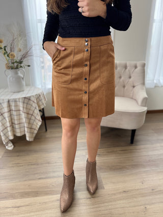 Person wearing a brown button-up skirt with a black top and brown boots in a room.