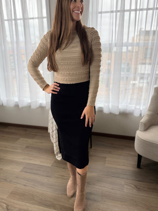 Woman wearing a beige ribbed sweater and black skirt indoors.