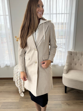 Woman wearing a beige coat in a room with curtains and furniture.