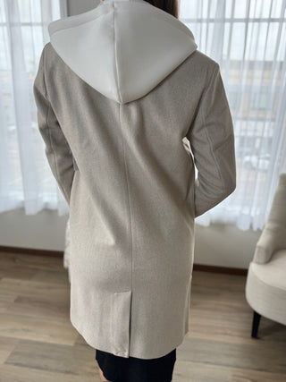 Person wearing a beige hooded coat in a room with white curtains.