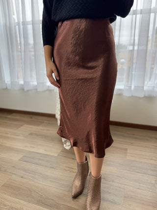 Person wearing a brown satin skirt and brown boots in a room with white curtains.