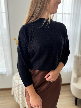 Woman wearing a black textured sweater and brown skirt in a room with white curtains.