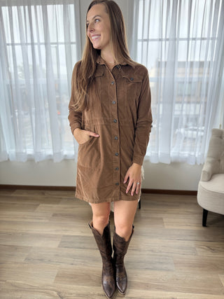 Woman wearing a brown dress and boots in a room with white curtains.