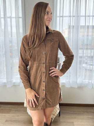 Woman wearing a brown corduroy dress standing in a room with white curtains.