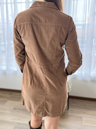 Person wearing a brown corduroy dress standing in a room with light-colored curtains.