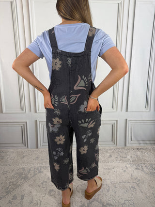 Person wearing floral overalls and a light blue shirt against a white paneled wall.