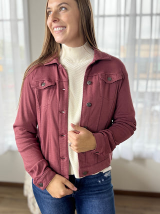 Woman wearing a maroon jacket over a white sweater and blue jeans indoors.
