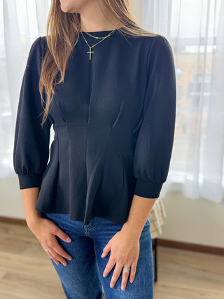 Person wearing a black blouse and blue jeans indoors with white curtains in the background