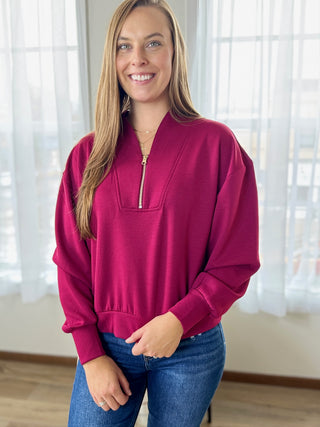 Woman wearing a magenta zip-up sweatshirt and blue jeans indoors.