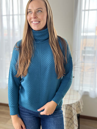 Woman wearing a blue turtleneck sweater standing indoors with a neutral background