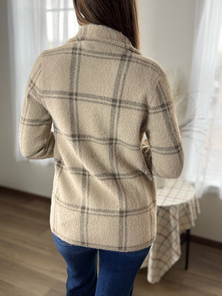 Person wearing a beige plaid jacket in a room with curtains and a table.