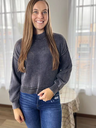 Woman wearing a dark gray sweater and blue jeans indoors.