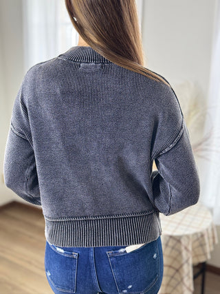 Person wearing a gray ribbed sweater and blue jeans in a neutral indoor setting