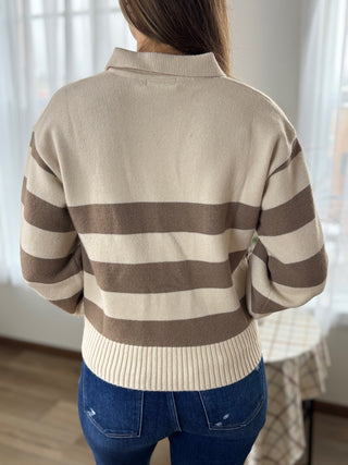 Person wearing a beige and brown striped sweater with blue jeans indoors.