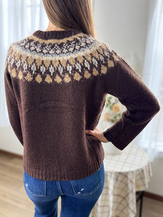 Person wearing a brown sweater with a patterned yoke in a room with curtains.