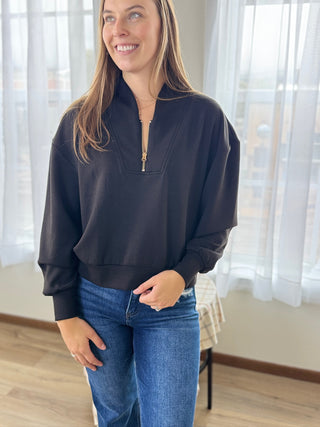 Woman wearing a dark blue zip-up sweatshirt and jeans indoors with light curtains in the background.