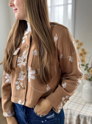 Woman wearing a brown jacket with floral embroidery in a blurred indoor setting