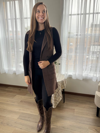 Woman wearing a long brown coat, black top, and brown knee-high boots in a room with large windows.