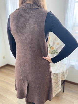 Person wearing a brown knitted vest over a dark long-sleeve shirt in a room with a table and chair.