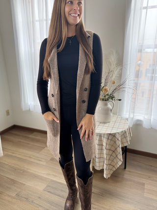 Woman wearing a long beige cardigan over a dark outfit in a room with curtains and a small table.