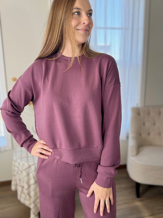 Woman wearing a mauve sweatshirt and pants in a room with a chair and window.