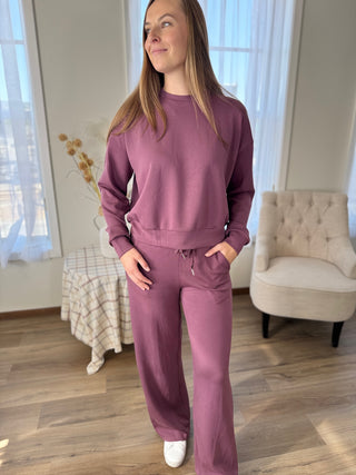 Woman wearing a purple tracksuit standing in a room with a chair and window.