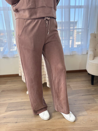 Person wearing brown tracksuit with white shoes in a room with wooden floor and white curtains.