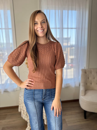 Woman wearing a brown sweater and blue jeans standing in a room with large windows.