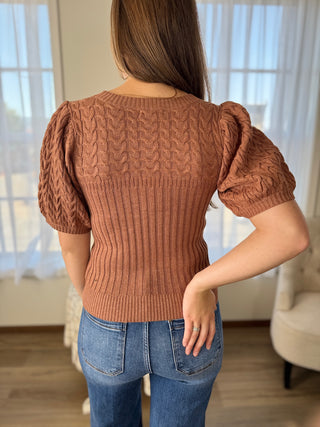 Woman wearing a brown cable knit sweater with puff sleeves indoors.