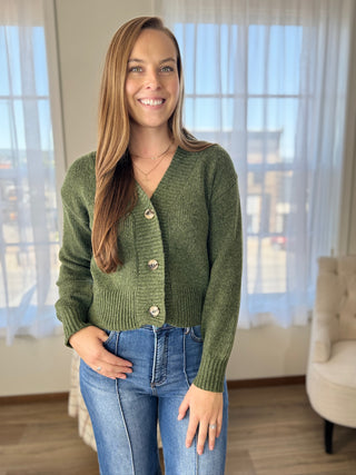 Woman wearing a green cardigan and blue jeans indoors with curtains in the background