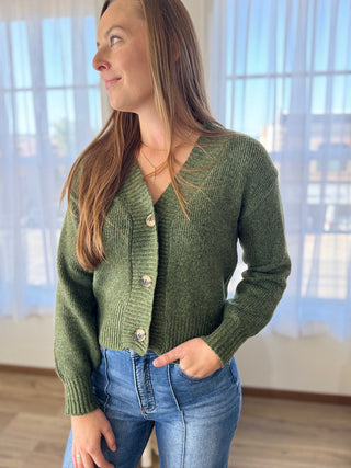 Woman wearing a green cardigan and blue jeans indoors with a blurred background