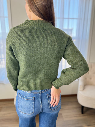 Person wearing a green sweater and blue jeans in a room with a window and chair.