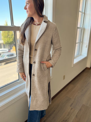 Woman wearing a beige checkered coat standing indoors near a window.