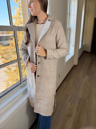 Woman wearing a checkered coat standing by a window with autumn foliage outside.