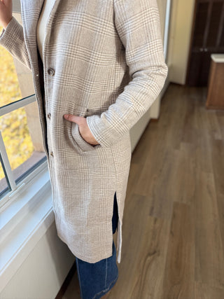 Person wearing a plaid coat standing indoors near a window with autumn leaves outside.