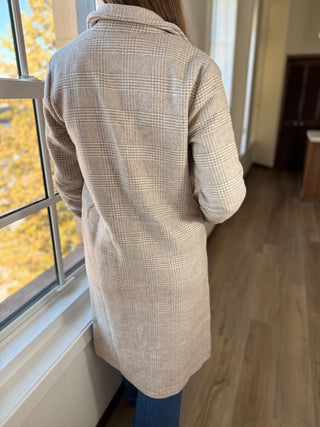 Person wearing a beige checkered coat standing by a window with autumn foliage outside.