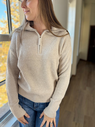 Woman wearing a beige sweater with a zipper, standing indoors near a window with autumn foliage outside.