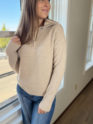 Woman wearing a beige sweater and blue jeans standing by a window.