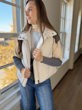 Woman wearing a beige puffer vest indoors with a window in the background
