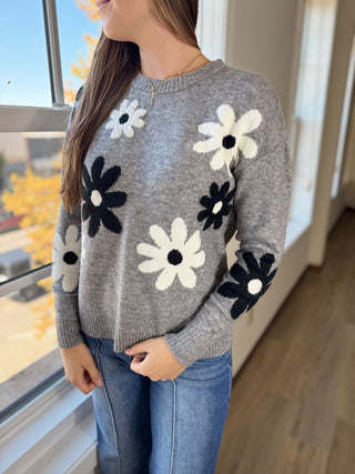 Person wearing a gray sweater with floral patterns standing indoors by a window.