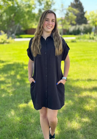 A woman standing in a grassy area wearing a black button-up dress with short sleeves, a collar, and front chest pockets.