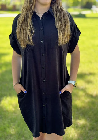 A woman standing in a grassy area wearing a black button-up dress with short sleeves, a collar, and front chest pockets.