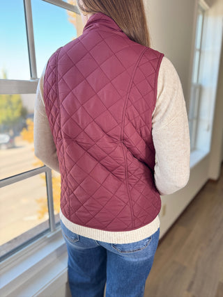Person wearing a maroon quilted vest indoors near a window with a view of trees outside.