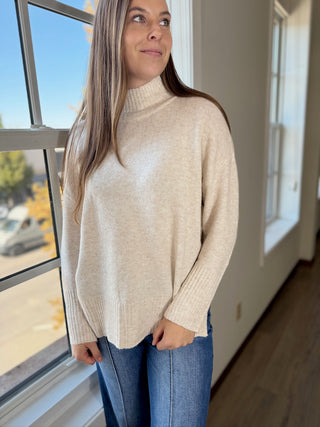 Woman wearing a beige turtleneck sweater and blue jeans standing by a window.