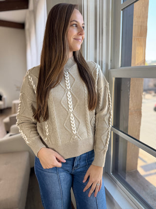 Woman wearing a beige cable knit sweater and blue jeans standing by a window.