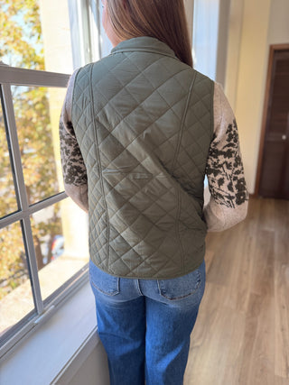 Person wearing a green quilted vest in a room with a window.