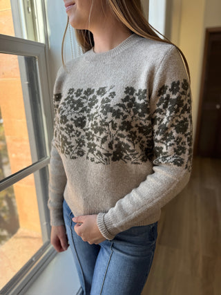 Woman wearing a beige sweater with black floral pattern standing by a window.