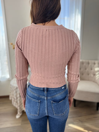 Person wearing a pink ribbed sweater and blue jeans in a room with light-colored walls and furniture.